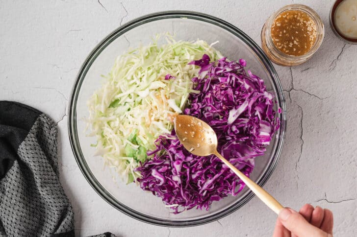 A glass bowl filled with green cabbage and purple cabbage, with a spoon drizzling sesame dressing over it.