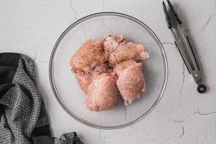 A glass bowl filled with raw chicken thighs sprinkled with seasonings.