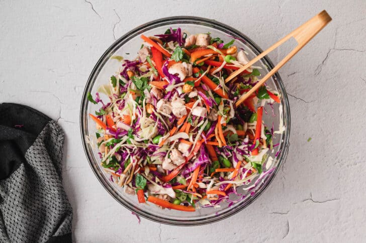 A glass bowl filled with chicken, red peppers, chow mein noodles, green onions, cilantro, carrots, cabbage, edamame and roasted peanuts, tossed together.