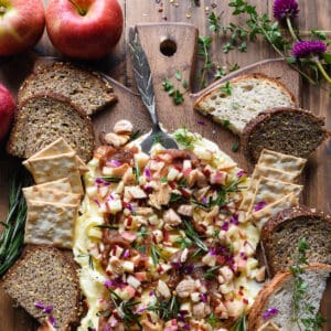 A butter board made with apple butter, fresh apples, nuts and herbs on a wooden cutting board, with bread and crackers for dipping.