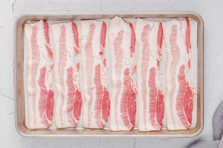 How to cook bacon in the oven, step one, which is lining up raw bacon on a rimmed baking pan.