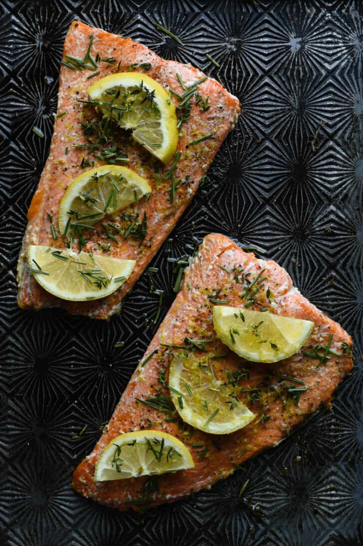 An oven baked salmon recipe, with fish covered in herbs and lemon slices on a textured metal baking pan.