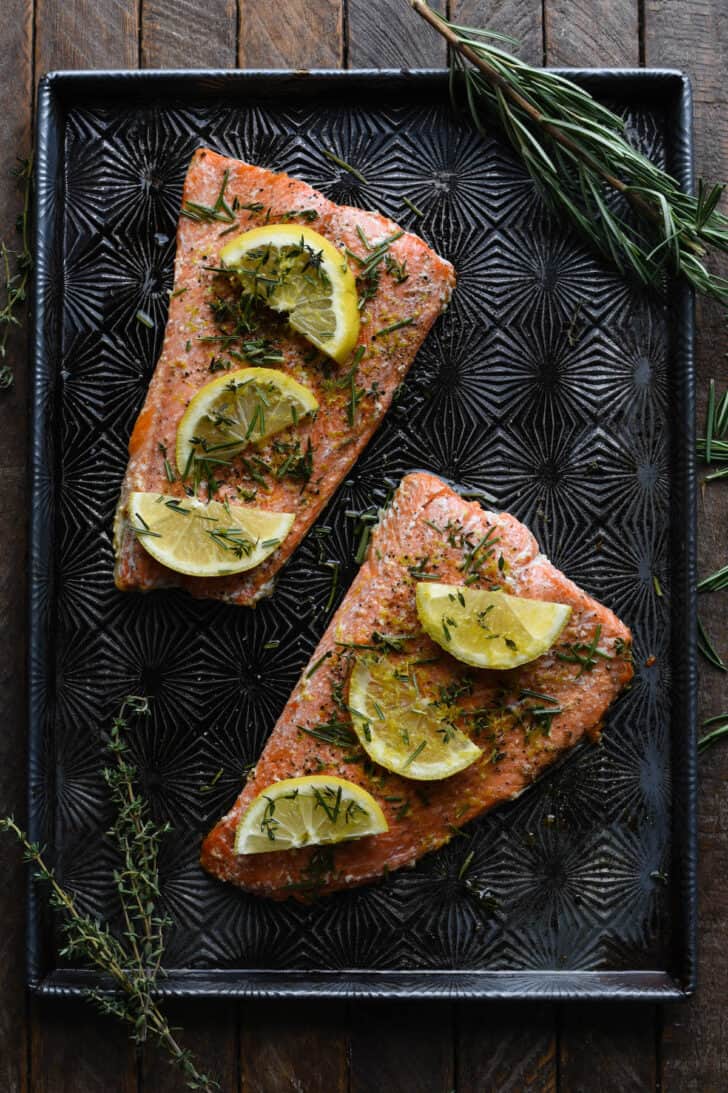 Oven baked salmon covered in herbs and lemon slices on a textured baking pan.