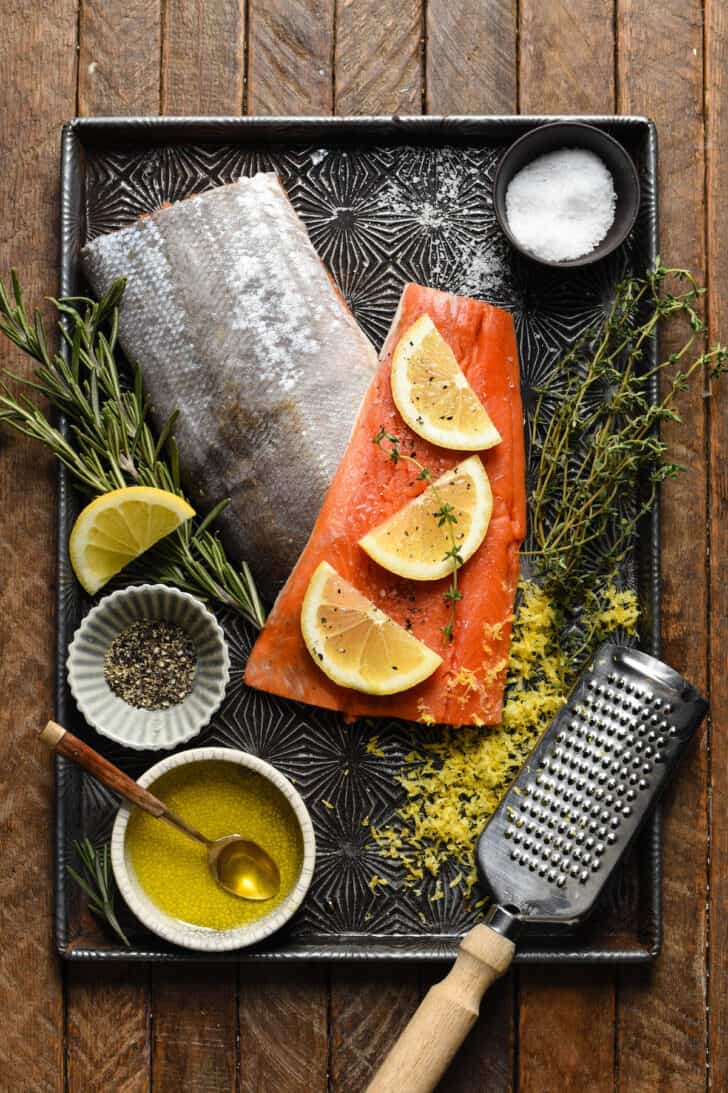 A textured baking pan filled with the ingredients including raw fish, lemon, herbs, salt and pepper.