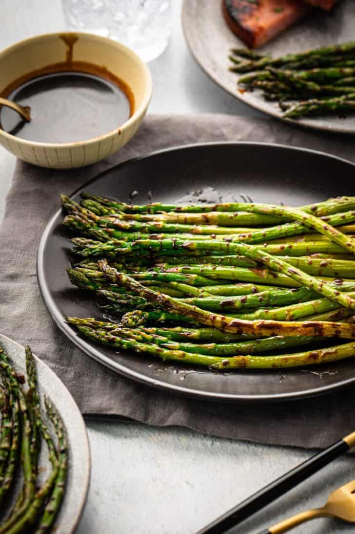 Oven roasted asparagus with balsamic glaze on a dark plate, with a small bowl of extra glaze in the background.