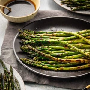 Roasted balsamic asparagus on a dark plate, with a small bowl of extra glaze in the background.