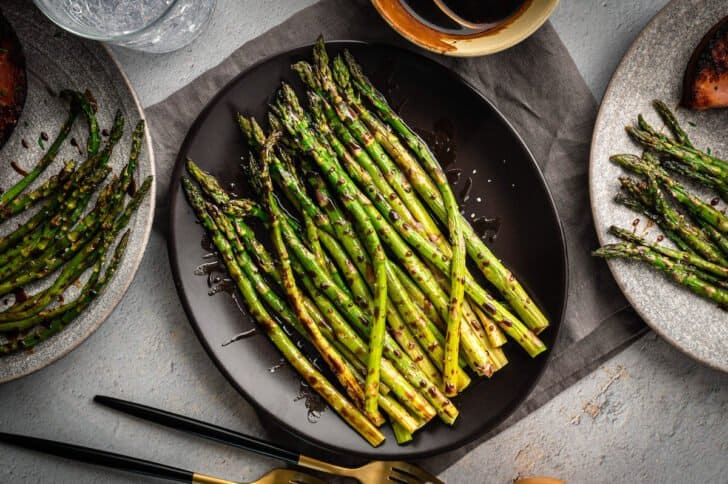 Roasted asparagus in the oven with balsamic glaze on a dark platter, with dinner plates nearby.