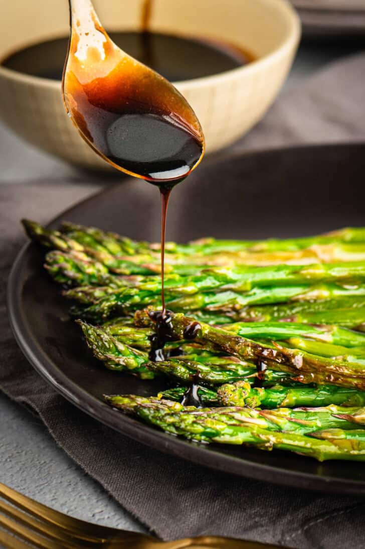 A gold spoon drizzling balsamic vinegar glaze over a bowl of roasted asparagus.