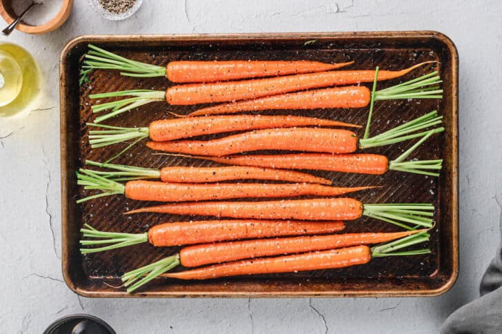 Whole raw root vegetables with some of their green tops attached, sprinkled with salt and pepper, on a rimmed baking pan.