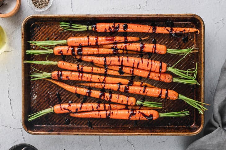 Whole roasted carrots with some of their green tops attached, on a rimmed baking pan, drizzled with balsamic glaze.