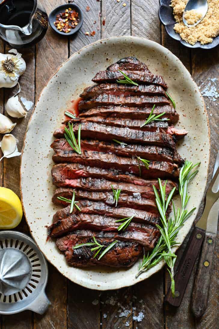 A cooked, sliced flank steak that has been marinated in steak marinade, on a speckled platter, with rosemary garnishing the steak.