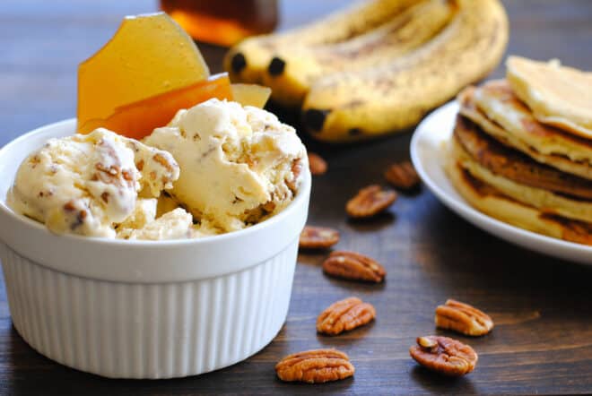 Large white ramekin filled with pancake ice cream, garnished with brittle. Bananas, pancakes and pecans garnish the scene.