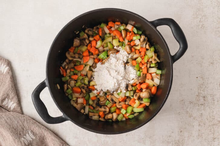 A black cast iron dutch oven filled with a mixture of mirepoix and mushrooms, with a bit of flour on top.