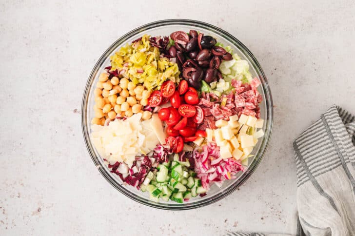 Chopped radicchio, lettuce, Provolone, salami, tomatoes, chickpeas, cucumber, onion, Parmesan, pepperoncini and olives in a glass bowl.