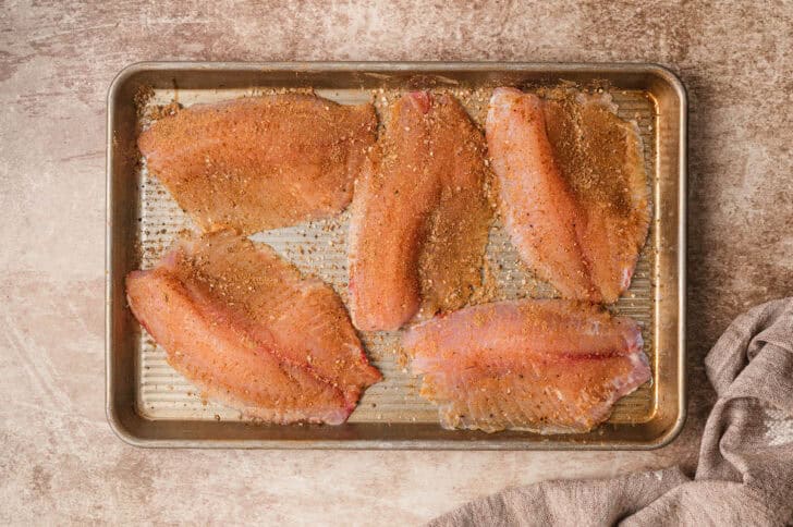 A rimmed baking pan topped with raw tilapia sprinkled with spices.