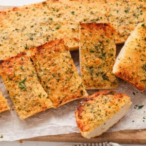 A loaf of homemade garlic bread on a wooden cutting board on a light surface.