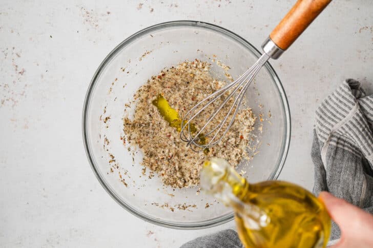 Olive oil being drizzled into a glass bowl with herbs and spices.