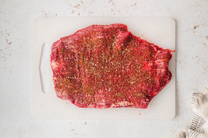 A piece of raw beef coated in seasonings on a white cutting board.