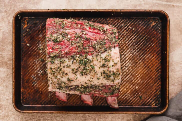 A rimmed baking pan topped with a raw standing rib roast rubbed in herbs.