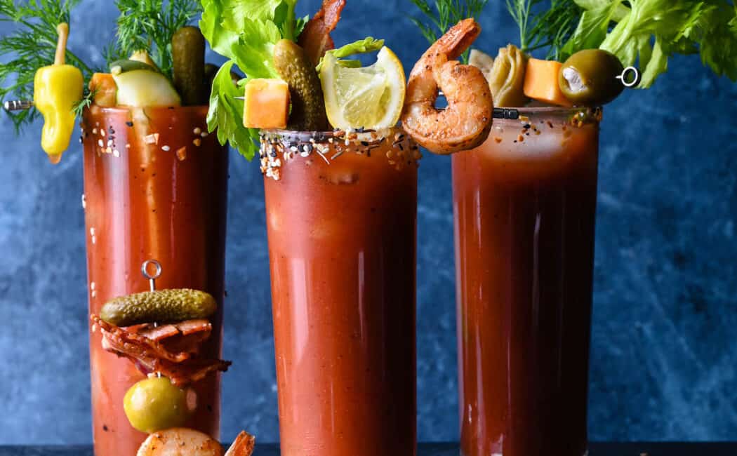 Three tall glasses filled with red juice, garnished with Bloody Mary skewers made with shrimp, bacon, cheese, pickles, artichokes and olives.