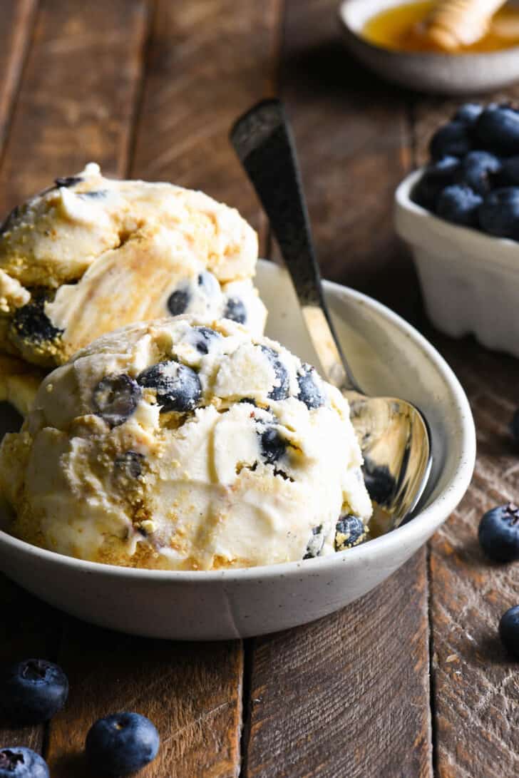 Small oval dish filled with scoops of blueberry cheesecake ice cream, with a bowl of blueberries and a dish of honey in the background.