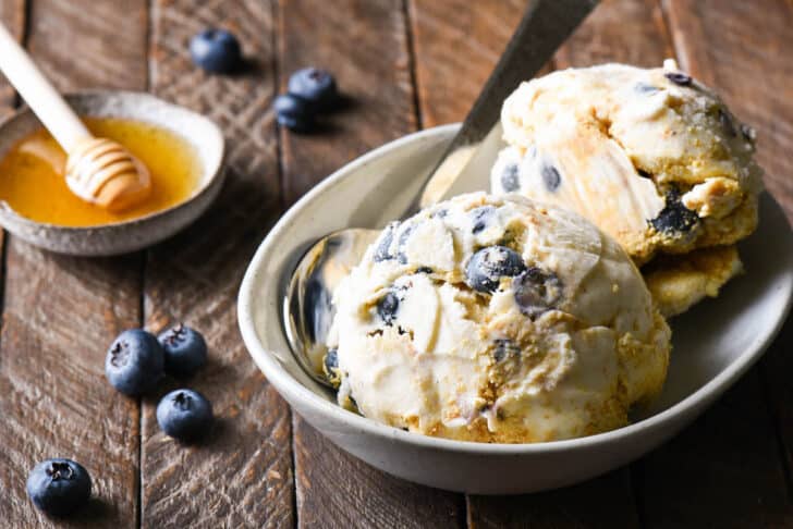 A small oval bowl filled with cream cheese ice cream swirled with graham cracker crumbs and blueberries, with blueberries and honey in the background of the scene.