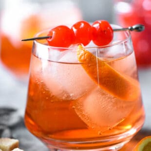 Orange hued cocktail with ice, orange slice and cherry skewer.