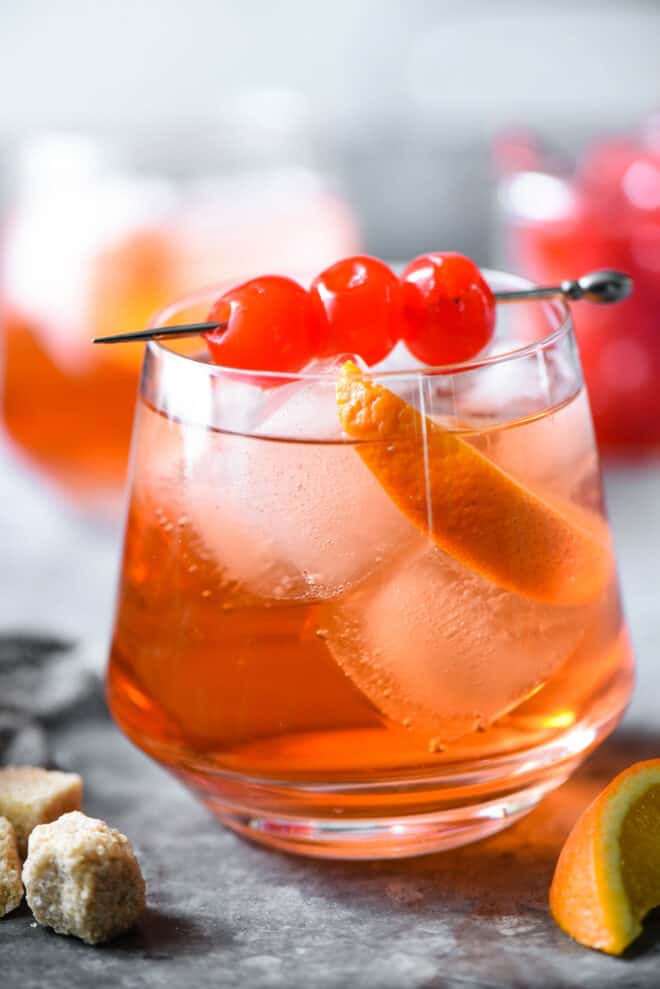 Orange hued cocktail with ice, orange slice and cherry skewer.