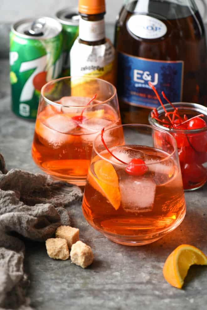 Two orange hued cocktails with maraschino cherries, brandy, orange bitters, and 7-up cans in background.