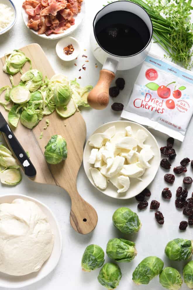 Ingredients for flatbread on a white surface, including cheese, bacon, root vegetables, herbs, dried cherries, and a pot of cherry glaze.