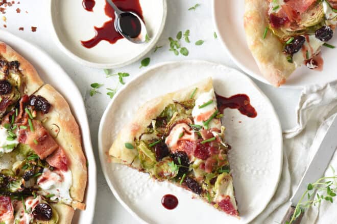 A tabletop scene of Brussels sprouts pizza being served onto small plates, with a cherry reduction sauce for drizzling.