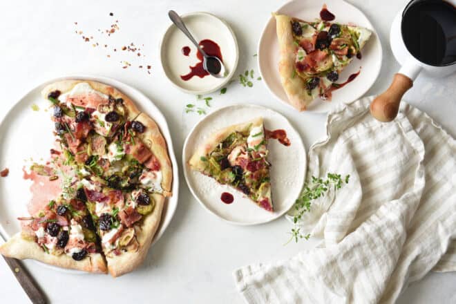 A tabletop scene of Brussels sprouts pizza being served onto small plates, with a cherry reduction sauce for drizzling.