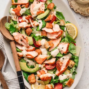 An oval platter filled with salmon Caesar salad with a wooden serving spoon.