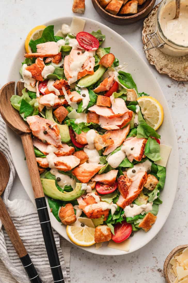 An oval platter filled with salmon Caesar salad with a wooden serving spoon.