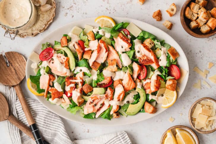 An oval platter filled with salmon Caesar salad with wooden serving spoons.