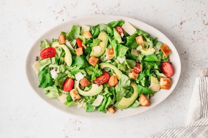 An oval platter topped with chopped lettuce, tomatoes, avocado, croutons, capers and shaved cheese.