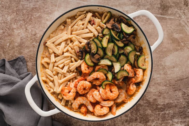 A white Dutch oven filled with a creamy orange sauce, casarecce pasta, cooked zucchini, mushrooms and shrimp, about to be stirred together.