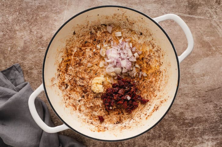 A white Dutch oven filled with shallots, garlic and sundried tomatoes.