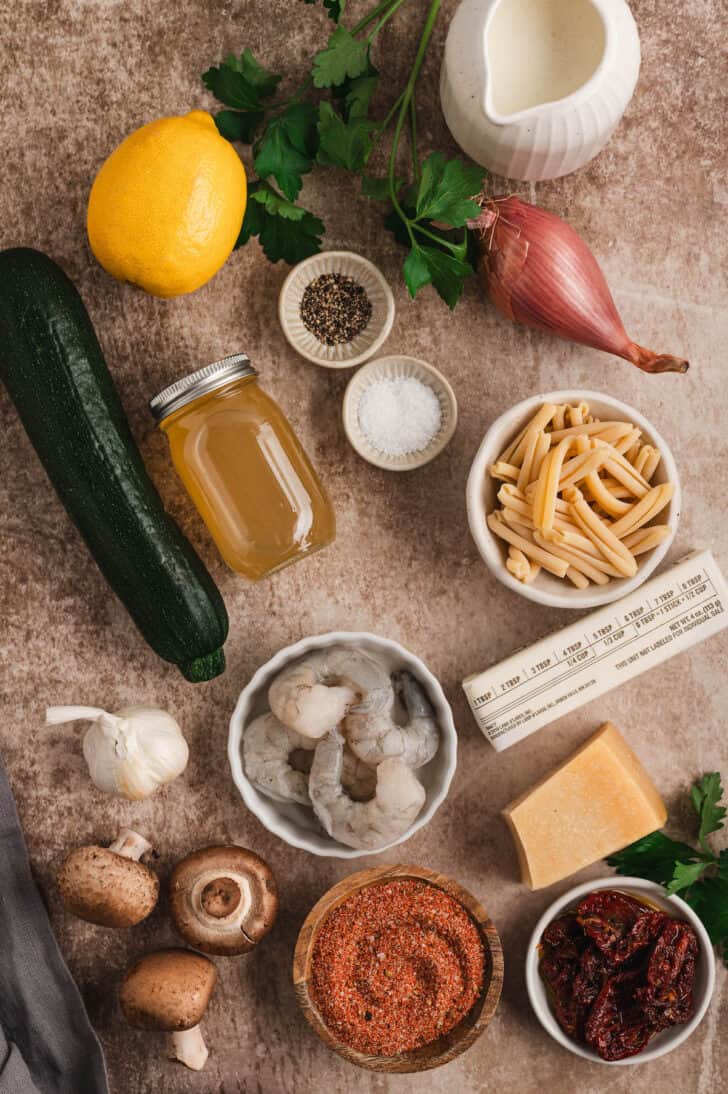 Ingredients on a textured brown surface, including dried noodles, spices, herbs, shallots, lemon, cream, butter, shrimp, mushrooms, tomatoes, cheese and garlic.