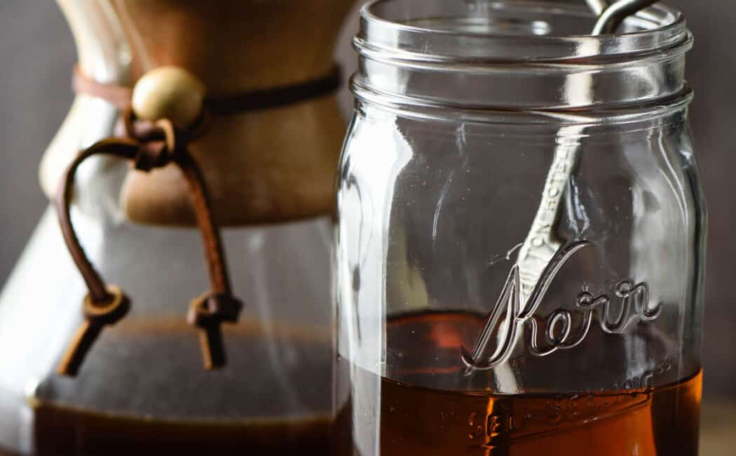 Quart sized mason jar half filled with brown syrup, with spoon in it. Chemex coffee maker is in background, caramel candies and coffee beans are in foreground.