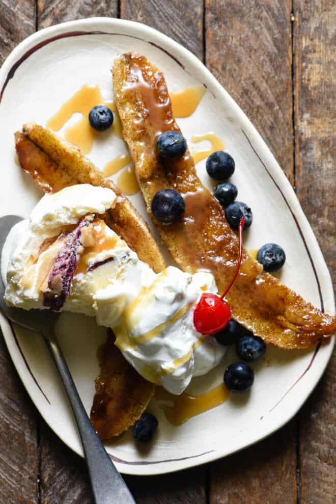 Small oblong shaped white plate topped with a caramelized banana, ice cream, blueberries, caramel sauce and a cherry.