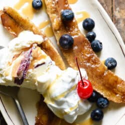 Small oblong shaped white plate topped with a caramelized banana, ice cream, blueberries, caramel sauce and a cherry.