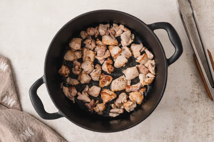 A black cast iron dutch oven filled with sauteed pieces of chicken thigh.