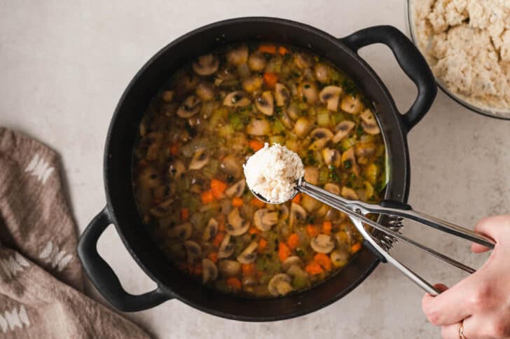 A hand scooping dumpling dough over a cast iron dutch oven filled with stew.