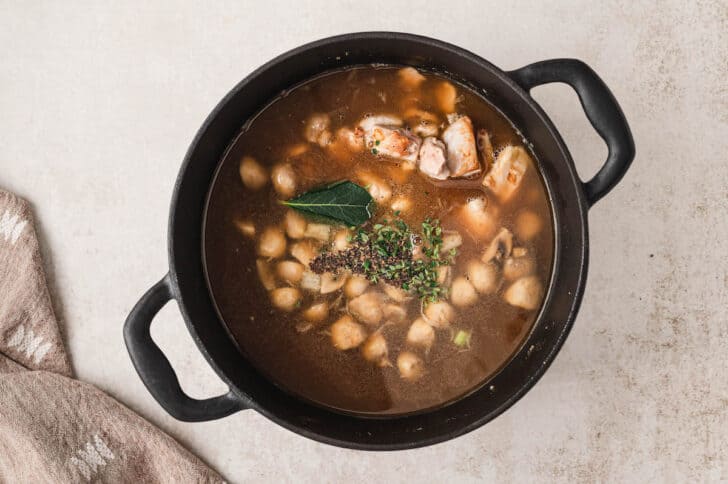 A black cast iron dutch oven filled with mushrooms, chicken, broth and herbs.