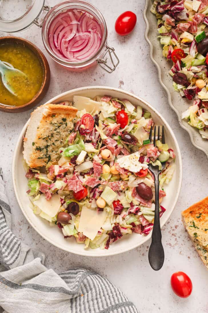 A scene showing an Italian chopped salad recipe on a large platter and a serving on a smaller plate, with garlic bread on the side.