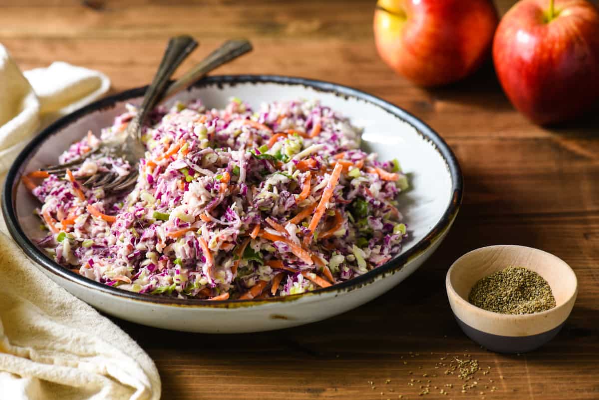 Shallow white bowl filled with coleslaw with celery seed, with smaller bowl of celery seed next to it, and apples in background.