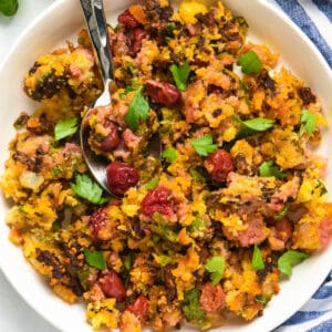 A white bowl filled with chorizo stuffing made with cornbread and white cherries, with a spoon digging into the stuffing.