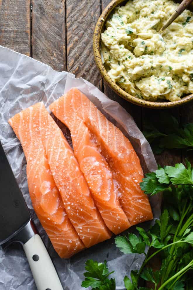 Rustic wood background with raw salmon fillets with slits cut down the center, and a bowl of crab filling.