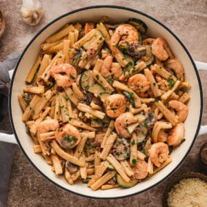 A white Dutch oven filled with Cajun shrimp pasta made with zucchini and mushrooms.